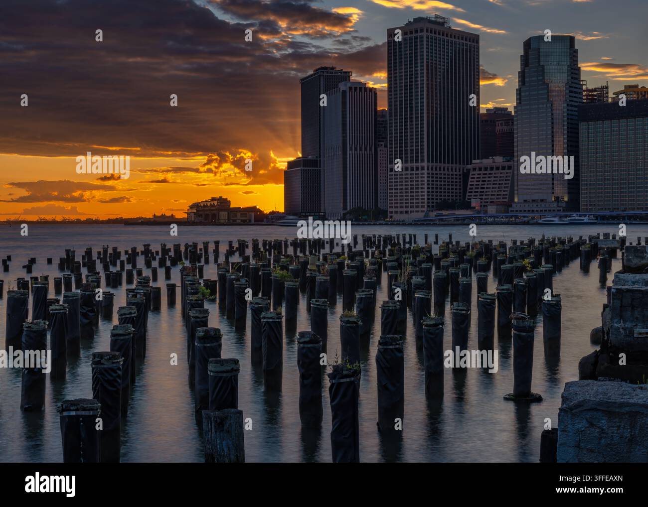 Die untergehende Sonne wirft ein warmes Licht über die Wolkenkratzer von Manhattan, wobei hölzerne Pfähle im Vordergrund faszinierende Silhouetten bilden. Wolken fügen hinzu Stockfoto