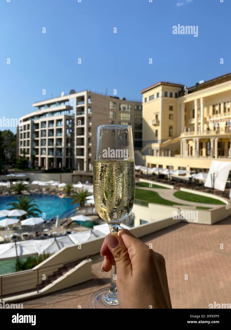 Hand hält funkelnden Wein über Resort-Pool im hellen Sonnenlicht. - Smartphone-aufgenommenes Stockfoto