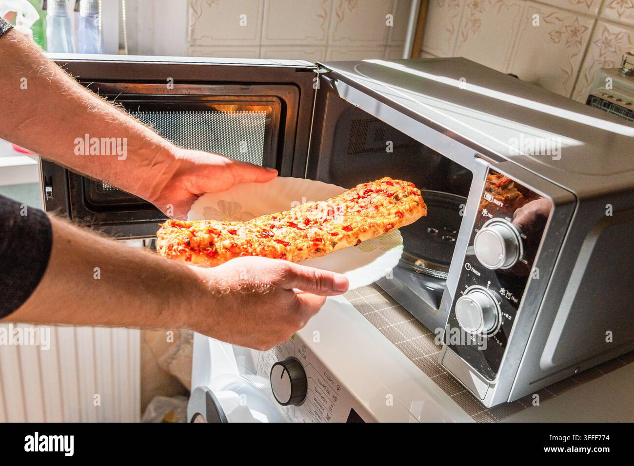 Hände, die eine gefrorene vegetarische Baguette-Pizza in eine Mikrowelle legen. Stockfoto