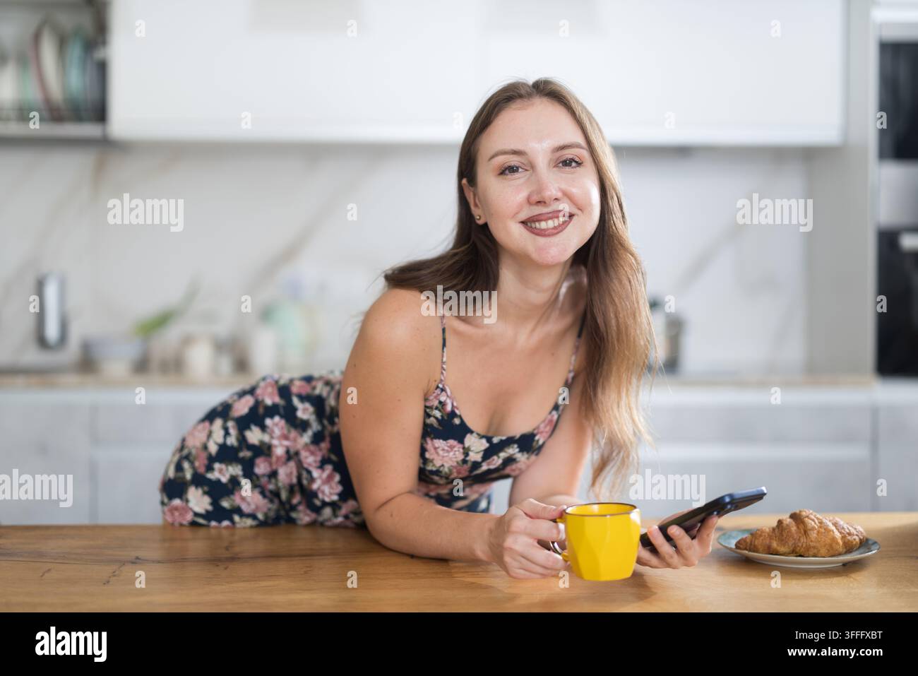Lächelnde Frau hat morgens eine Tasse heißen Kaffee gemacht, sie hält ein Mobiltelefon in den Händen und scrollt durch soziale Netzwerke. Frau, die eine SMS schreibt Stockfoto