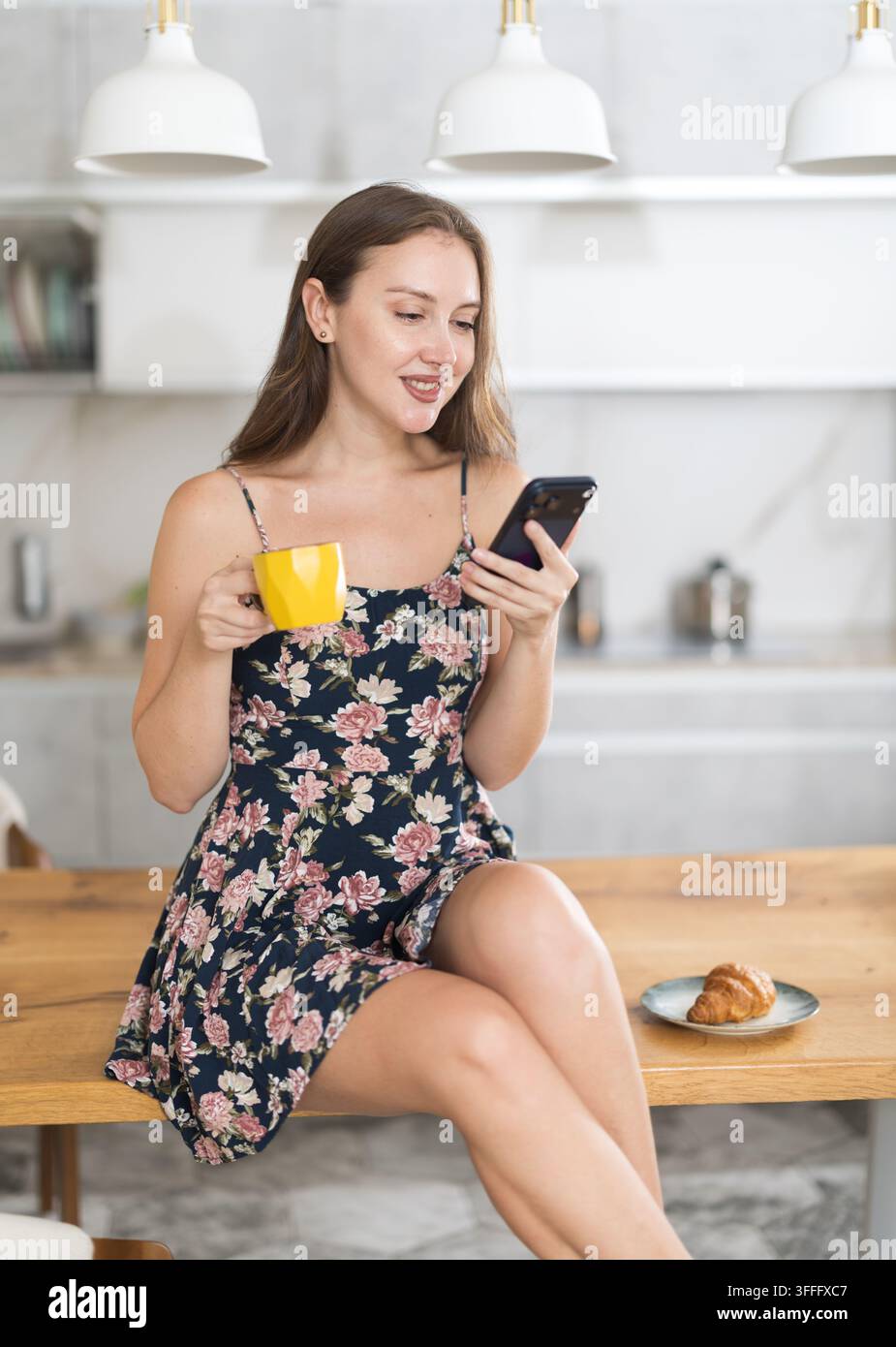 Lächelnde Frau hat morgens eine Tasse heißen Kaffee gemacht, sie hält ein Mobiltelefon in den Händen und scrollt durch soziale Netzwerke. Frau, die eine SMS schreibt Stockfoto