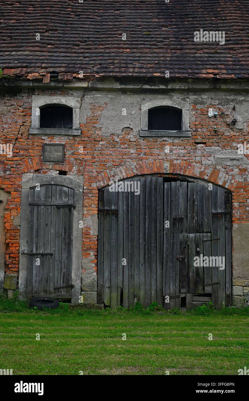 Alte Backstein-Scheune mit historischem Holztor und Herrenhaustüren unter einem verwitterten Ziegeldach – historische ländliche Architektur in der Region Złotoryja. Stockfoto