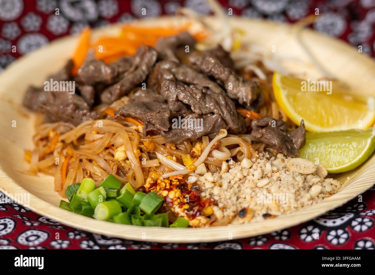 Beef Pad Thai, ein beliebtes thailändisches geröstetes Reisnudelgericht, garniert mit geschnittenem Rindfleisch, Limette, zerkleinerten Erdnüssen und Frühlingszwiebeln, serviert in einer Bambusschale Stockfoto