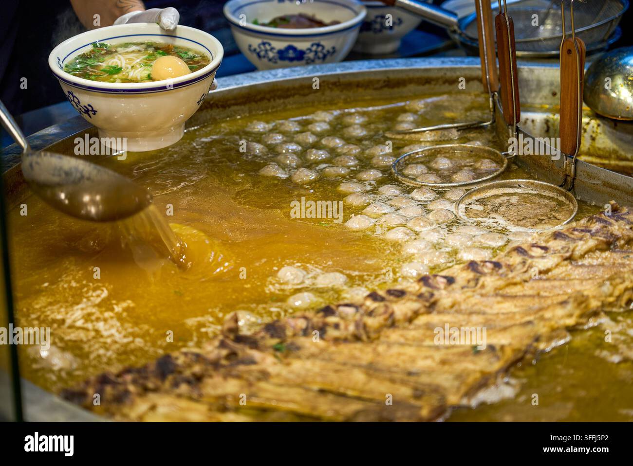 Eine große blau-weiße Porzellanschale enthält Guizhou-Rindernudeln, geschmorte Eier und Gemüse, während Dampf von einem großen Topf mit Brühe hinter Ihnen aufsteigt. Stockfoto