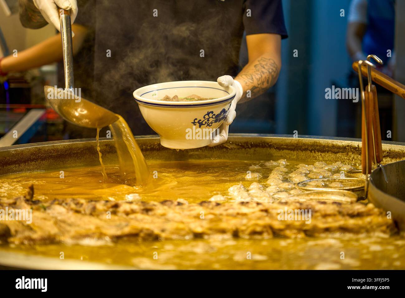 Mit Handschuhen schöpft der Besitzer des Guizhou-Rindernudelessens goldene Rindfleischsuppe aus einem großen Topf mit einem großen Löffel. Stockfoto