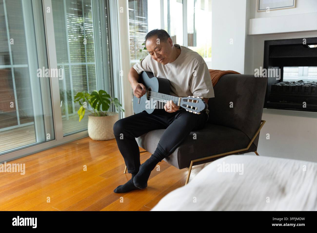 Seniorer asiatischer Mann sitzt auf dunklem grauem Sessel und spielt schwarze klassische Gitarre in einer Schlafzimmerecke Stockfoto