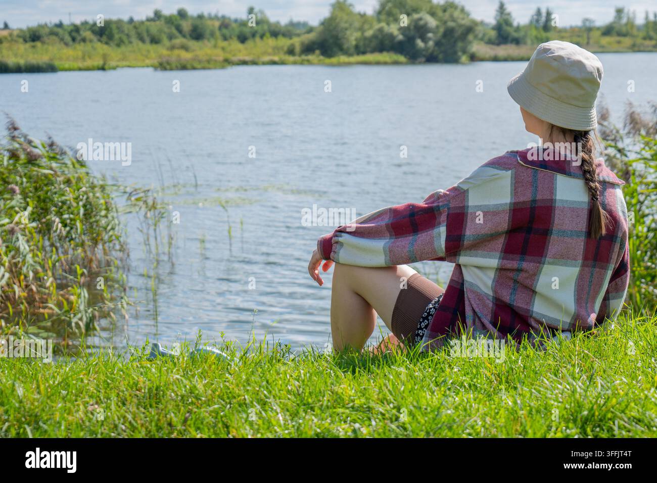 Frau mit einem leichten Sonnenhut und kariertem Hemd sitzt seitlich auf hellem grünem Gras neben dem Wasser mit untergeschlagenen Knien. Sie blickt in Richtung eines f Stockfoto