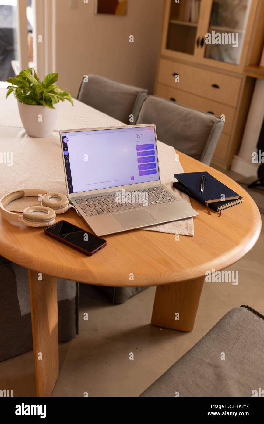 Offener silberner Laptop zeigt eine Chat-Schnittstelle auf einem runden Tisch zu Hause mit Kopfhörern und Telefon Stockfoto