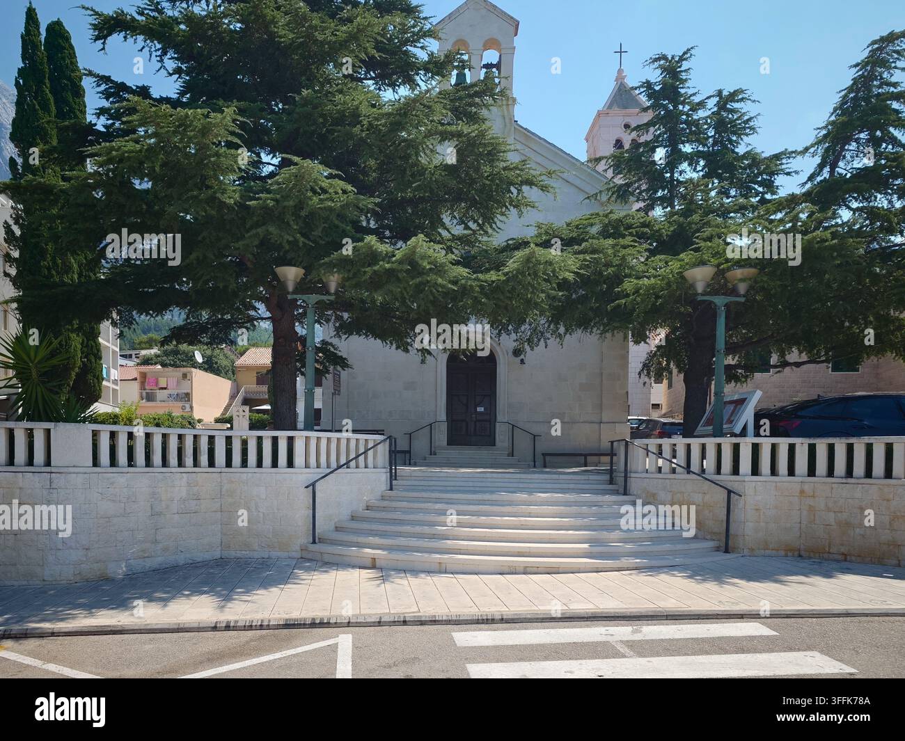 Ein malerischer Blick auf die St. Nikole Kirche mit einer Steinfassade, hohen Zypressen und einem Fußgängerüberweg bei hellem Tageslicht. - Smartphone-aufgenommenes Stockfoto