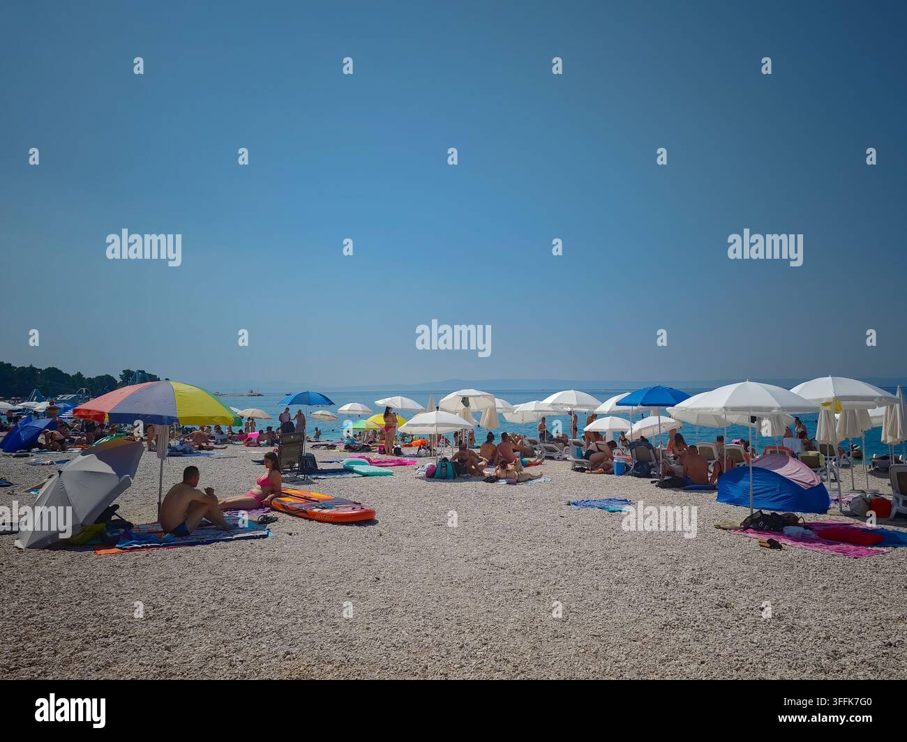 Überfüllter Kieselstrand unter einem klaren blauen Himmel am Nikolina-Strand in Baska Voda mit bunten Sonnenschirmen und entspannten Touristen. - Smartphone-aufgenommenes Stockfoto