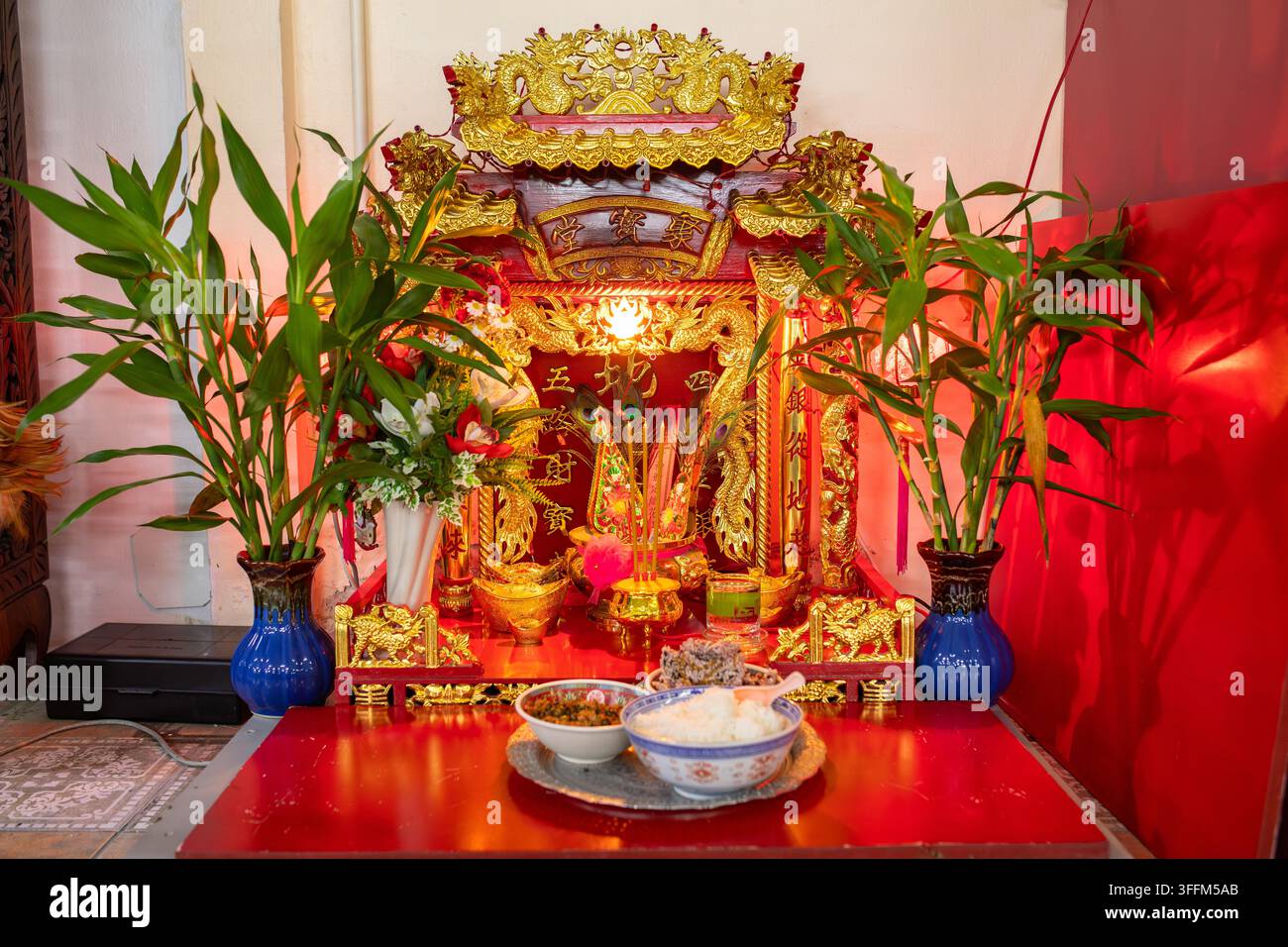Goldener Geisterhaus-Schrein leuchtet sanft im Inneren, umgeben von grünen Pflanzen und kleinen Opfergaben, rote Innenwand verleiht Wärme, während Kerzenlicht schafft Stockfoto