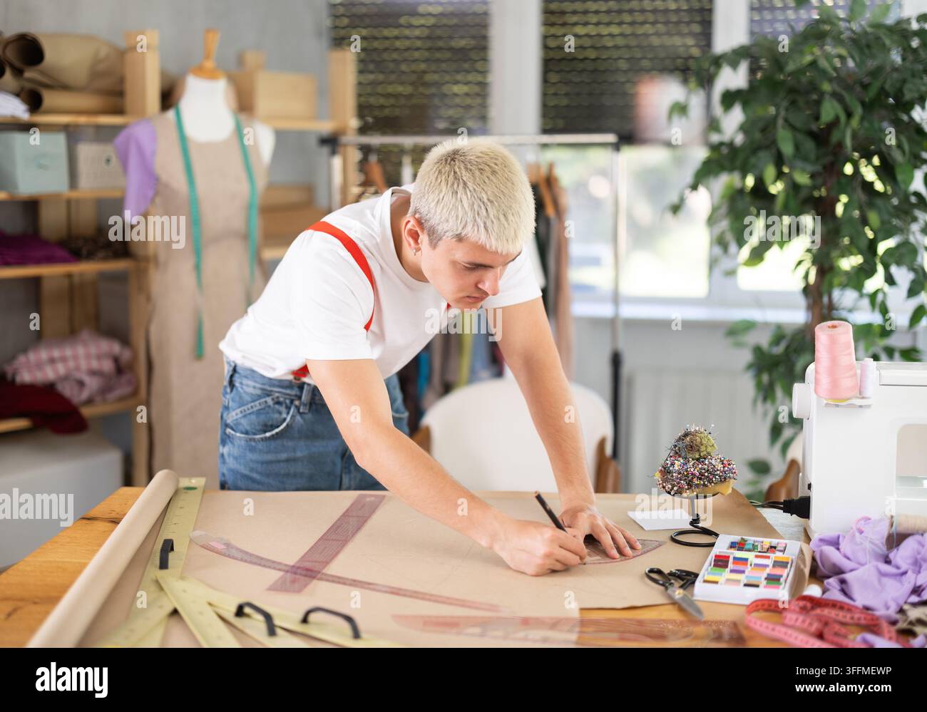Junger männlicher Schneider zeichnet ein Muster auf ein Blatt Papier mit Kurven, während er ein Kleid an seinem Arbeitsplatz in einer Mode-Nähwerkstatt näht Stockfoto