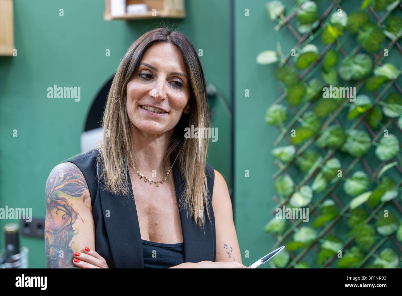 Professionelle Friseurin mit tätowiertem Arm lächelt und hält eine Schere Stockfoto