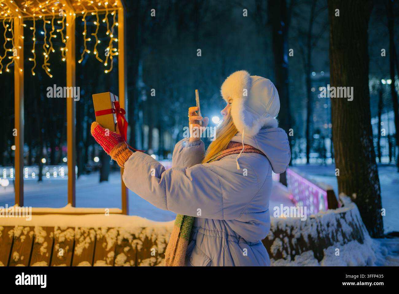 Frau steht in einem verschneiten Winterpark in der Nacht. Hält ein eingewickeltes Weihnachtsgeschenk. Nutzt ihr Handy, um unter dem warmen Schein dekorativer Lichterketten ein Selfie zu machen. Fängt die Feiertagsstimmung ein und teilt sie Stockfoto