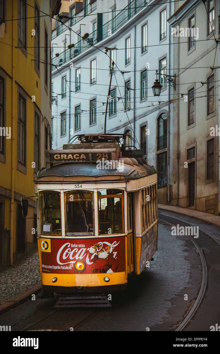 Historische Straßenbahn 28E fährt durch eine enge Straße in Lissabon, Portugal, zur Graça, mit traditionellem gelbem Straßenbahnwagen und Oberleitungen. Stockfoto