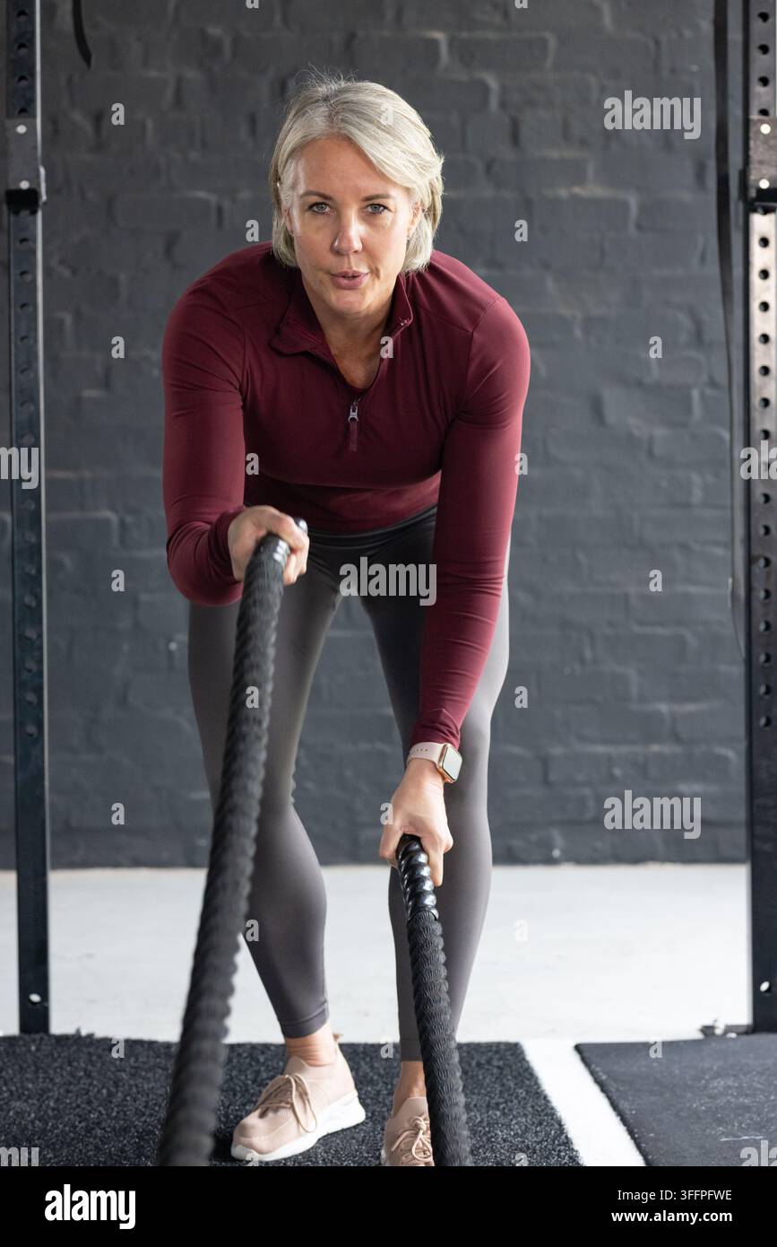 Seniorin führt ein Battle-Rope-Training auf einer Gummimatte im Fitnessstudio durch, trägt ein kastanienbraunes Reißverschluss-Top Stockfoto