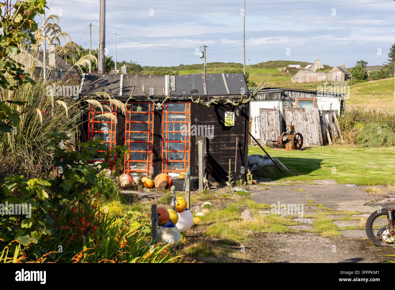 Schwarzer Schuppen mit orangefarbenen Fensterrahmen und Geweihen, umgeben von Bojen und überwucherten Pflanzen an den Whaligoe Steps, Schottland, UK. Stockfoto