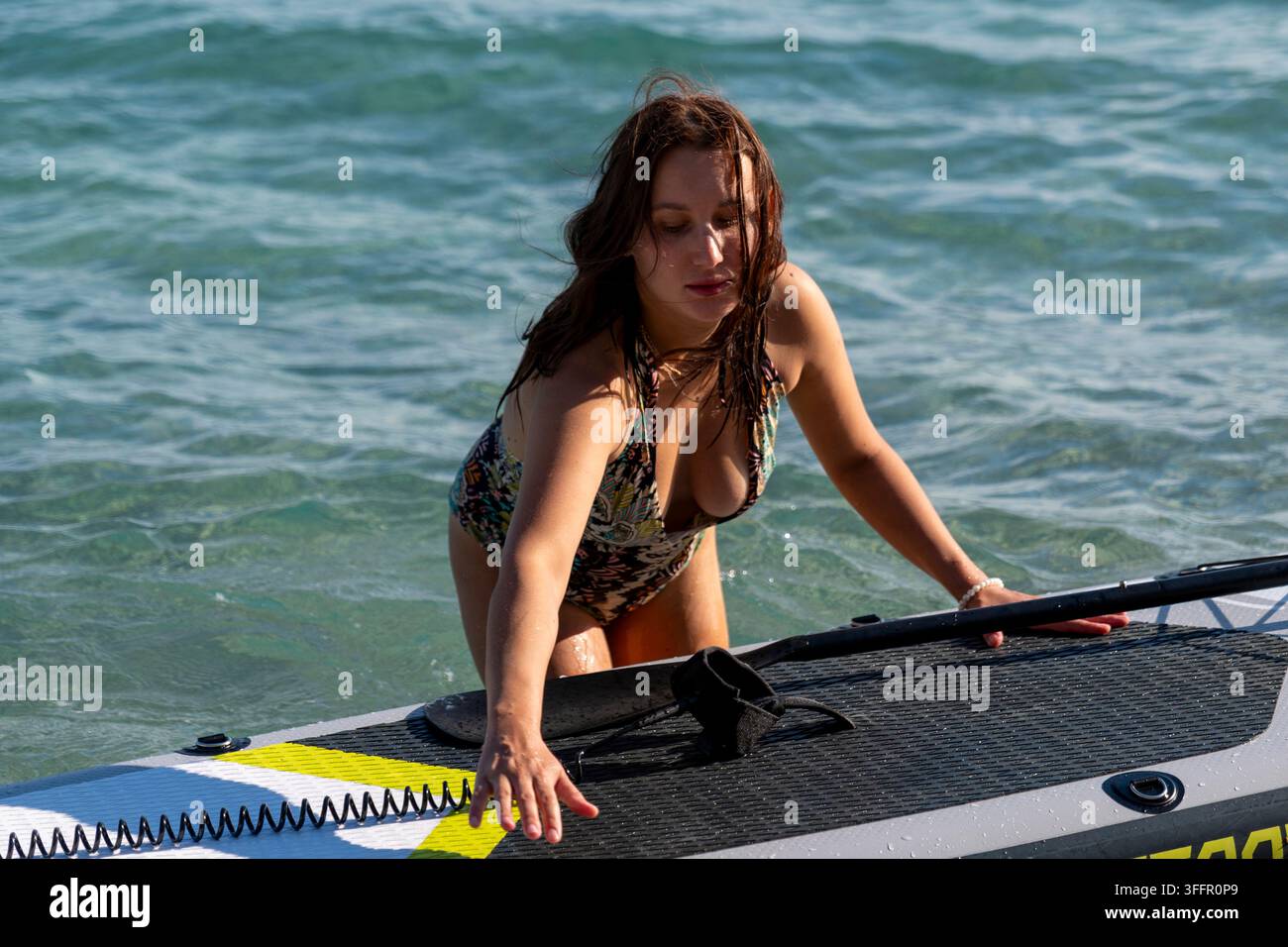 Eine junge Frau in einem Badeanzug steigt in klarem Ozeanwasser auf ein Paddelboard. Stockfoto
