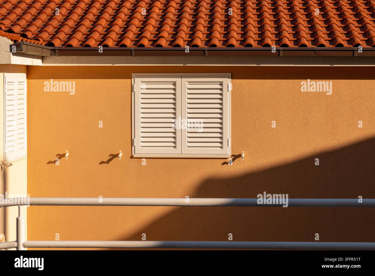 Eine orangefarbene Stuckhauswand mit geschlossenen weißen Fensterläden und starken diagonalen Schatten. Stockfoto