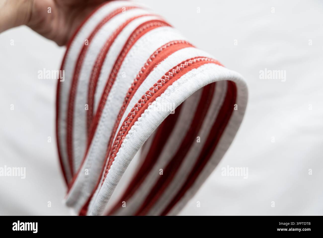 Rot-weiß gestreiftes Stoff-Stirnband, das von der Hand gehalten wird, zeigt genähte Kanten, gewebte Textur und gebogene flexible Form vor hellem Hintergrund. Stockfoto
