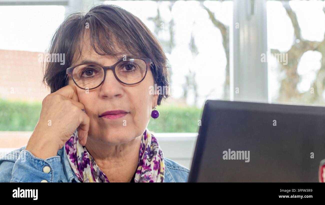 Nahaufnahme-Porträt einer ernsten Frau mittleren Alters mit Brille und einem bunten Schal, die sich neben einem Laptop auf ihre Hand stützt und remote arbeitet Stockfoto