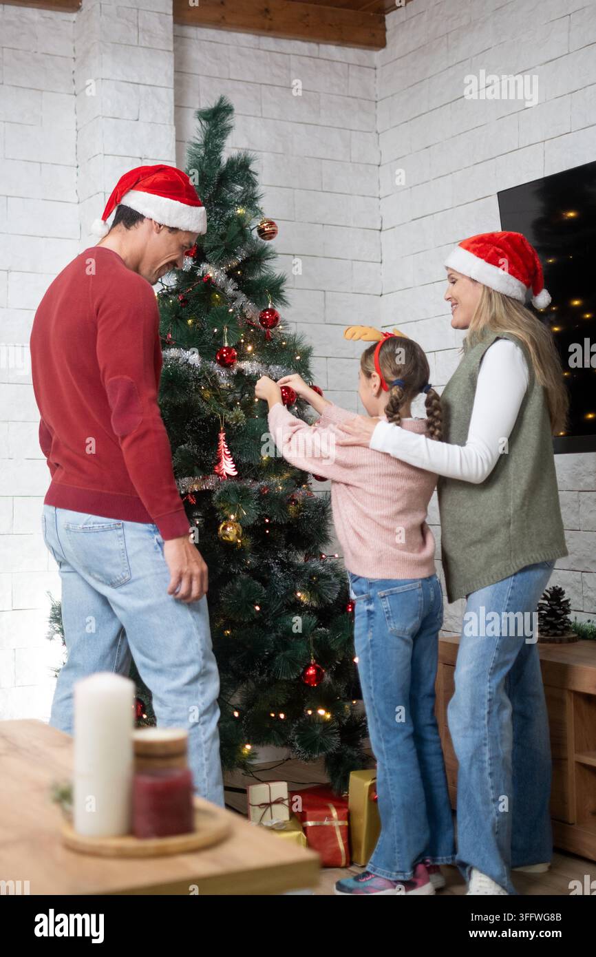 Eltern und Tochter schmücken gemeinsam einen Weihnachtsbaum, während sie festliche Weihnachtsmannmützen während der Feiertagssaison tragen. Stockfoto