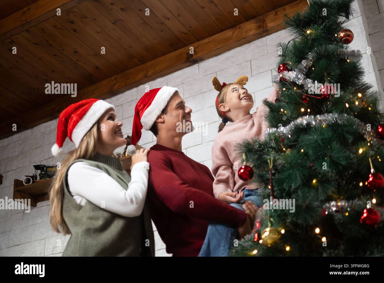 Eltern und Tochter schmücken gemeinsam einen Weihnachtsbaum, während sie festliche Weihnachtsmannmützen während der Feiertagssaison tragen. Stockfoto