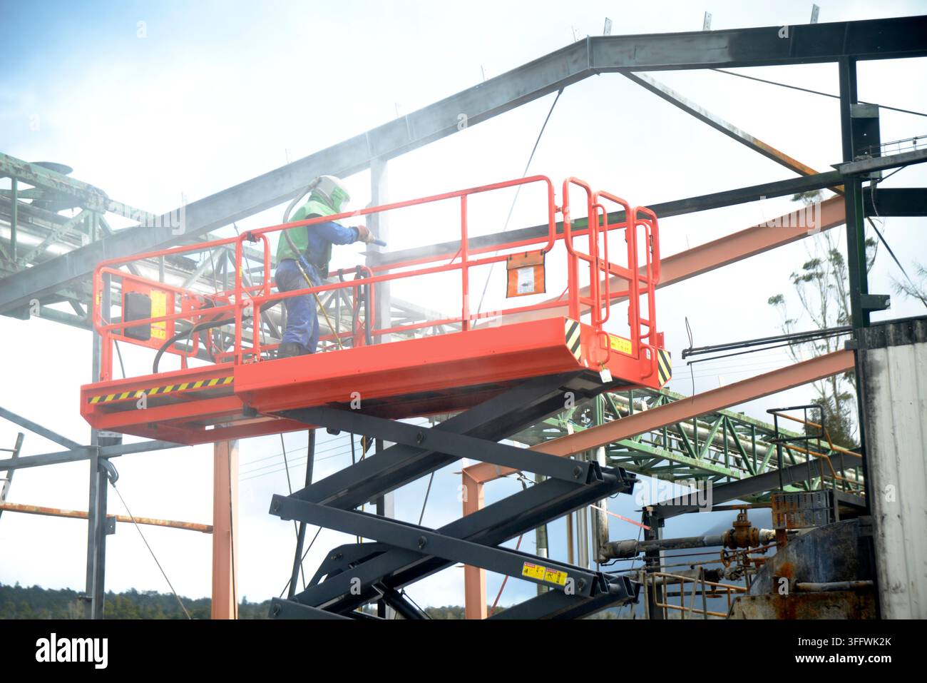 Ein Mann in Schutzkleidung bedient einen Sandstrahler von einer Scherenbühne aus, um Stahlarbeiten an einem feuerbeschädigten Gebäude zu reinigen, bereit für die Neubemalung. Stockfoto