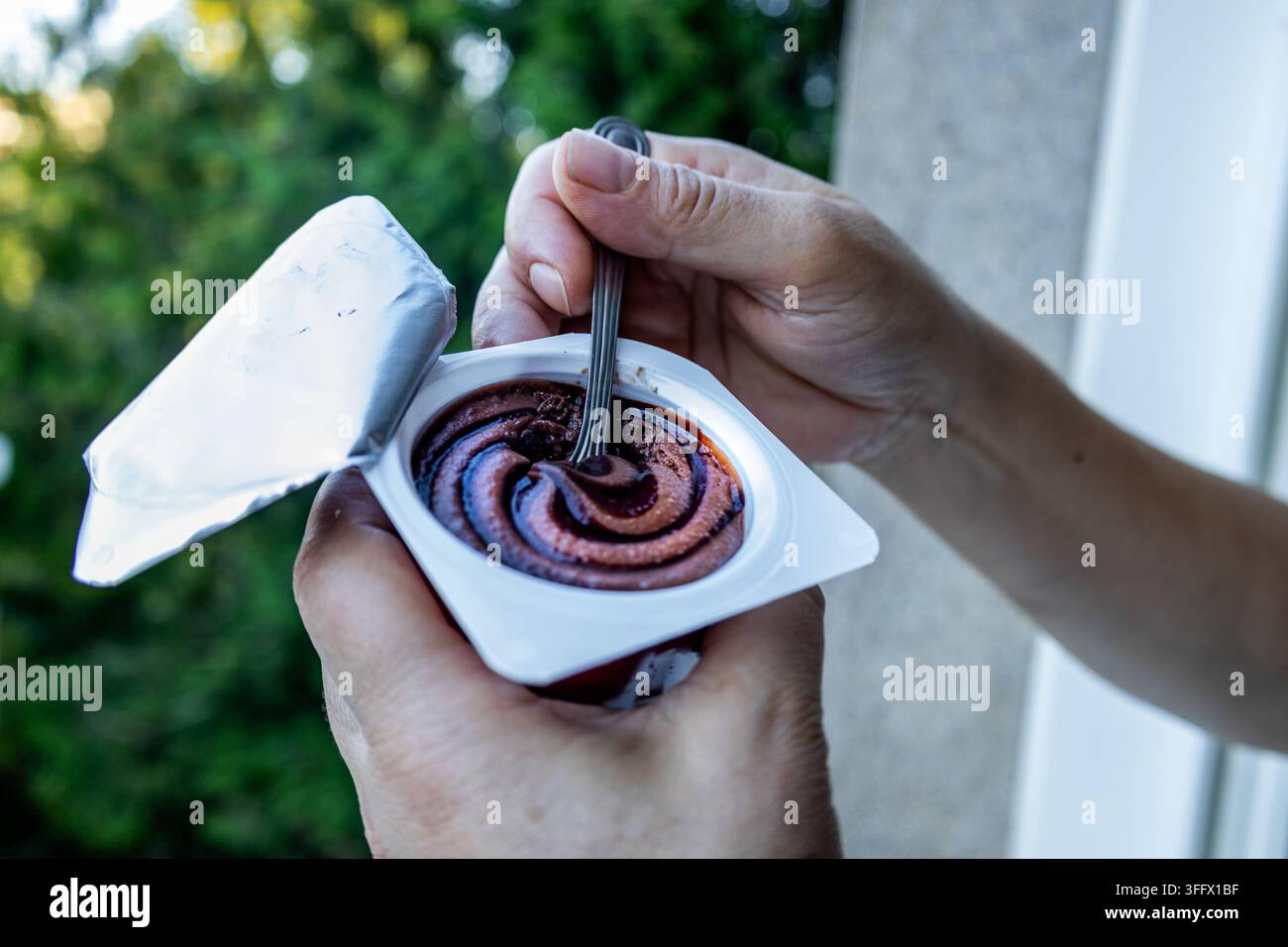 Hände halten eine offene Tasse mit cremigem Schokoladendessert und einem Löffel und zeigen eine üppige Spirale im Freien vor weichem grünem Laub Stockfoto