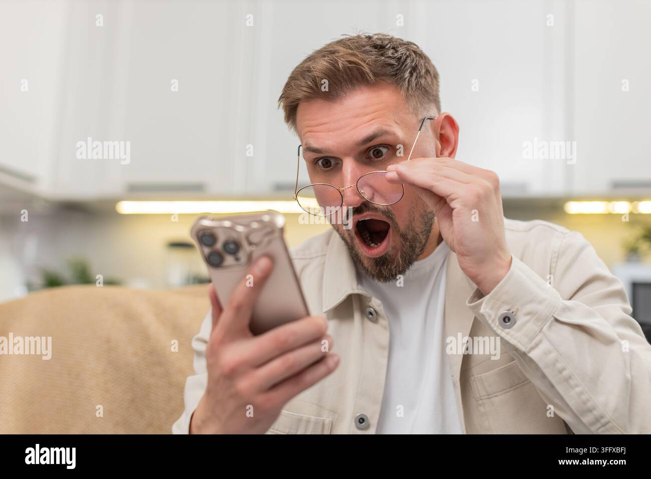 Ein junger Mann zu Hause liest mit einer Brille winzige Texte auf dem Smartphone und staunt über die Geschichte und Neuigkeiten. Der kaukasische Kerl auf dem Sofa sieht starke Aufmerksamkeit im Boten, zeigt Freude Erleichterung und helle Wow-Überraschung auf einmal Stockfoto