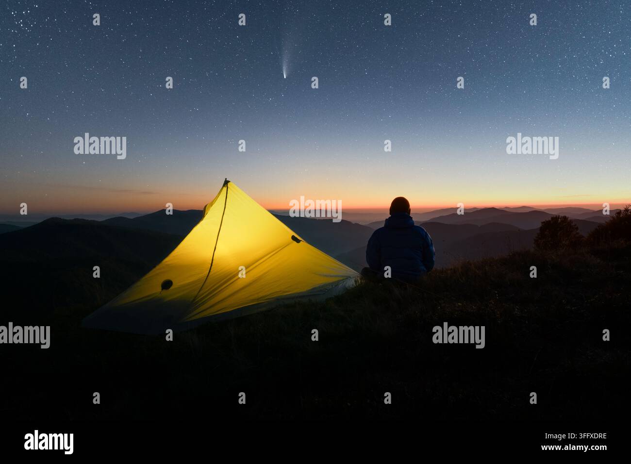 Gelbes Zelt und ein Wanderer, der auf einem Bergkamm unter einem sternenklarem Nachthimmel ruht. Schwache Aurora und ferne Berge schaffen eine friedliche Wildnis-Camping-Szene Stockfoto