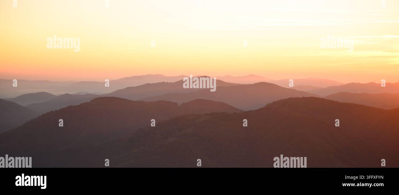 Schichtige Bergkämme glühen im warmen Licht bei Sonnenaufgang mit einem dunstigen goldenen Himmel. Minimale Panoramalandschaft mit atmosphärischen fernen Gipfeln Stockfoto
