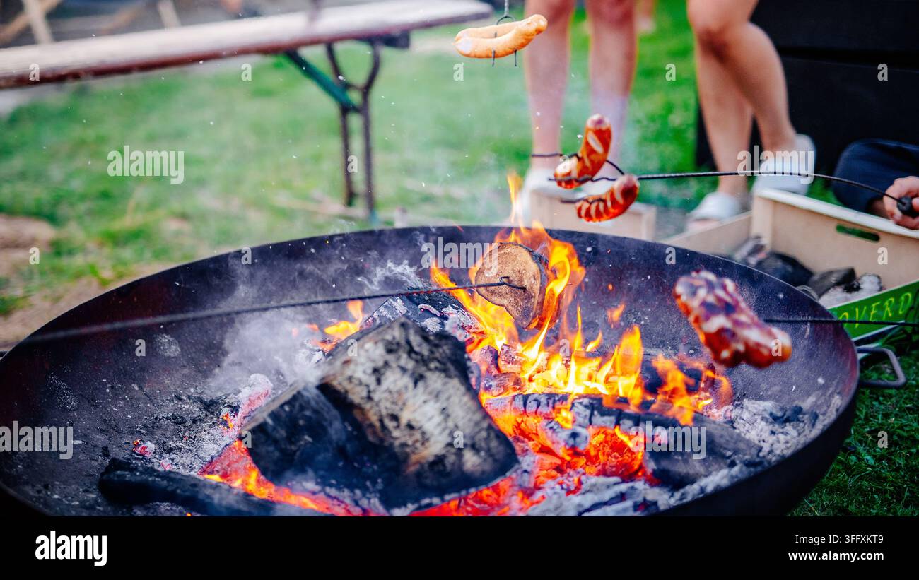 Freunde versammeln sich um einen Feuerkorb und grillen Würste auf Spießen während einer Abendparty Stockfoto