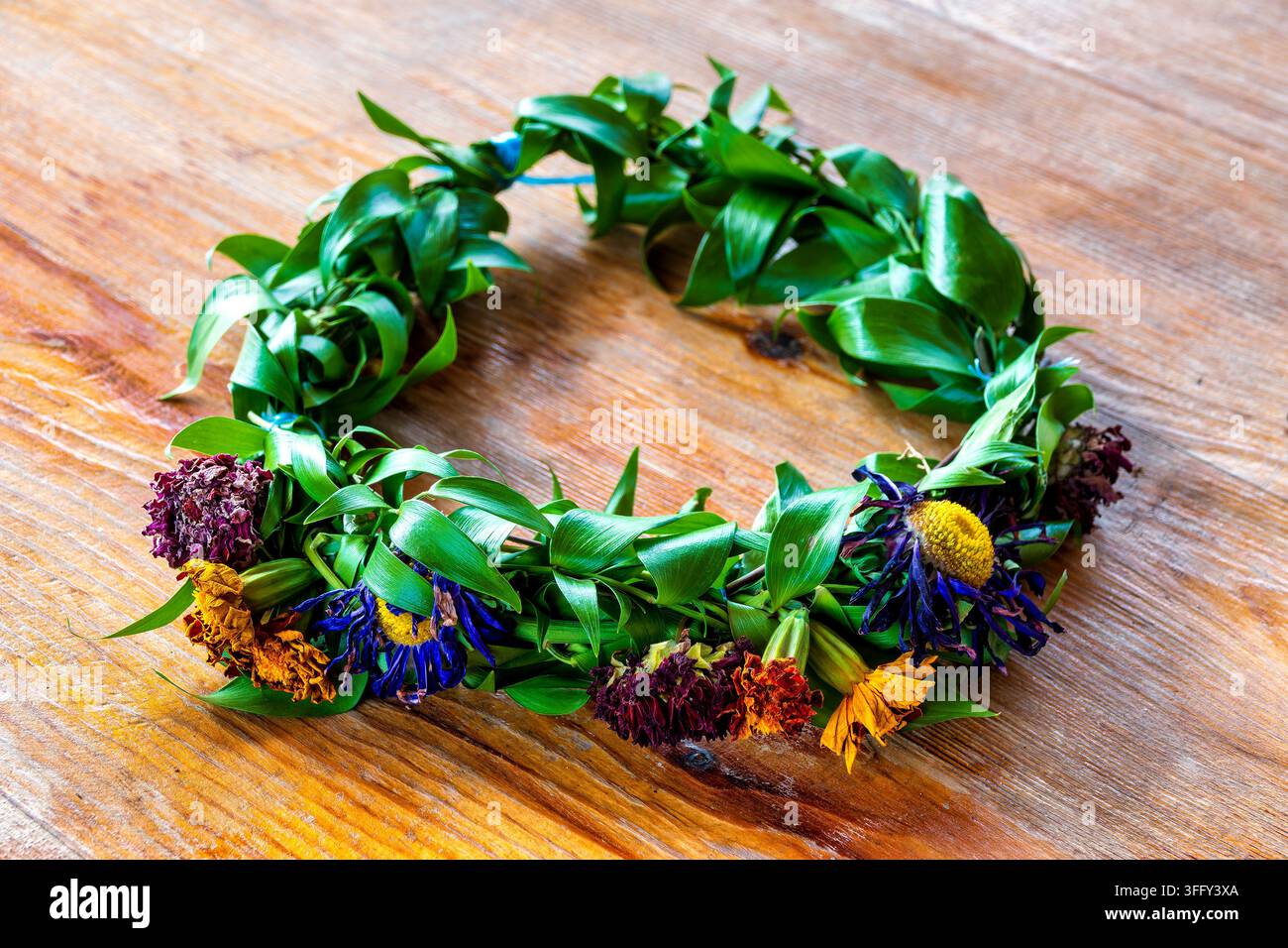 Handgefertigter Kranz mit grünen Blättern und bunten getrockneten Blumen auf einem rustikalen Holztisch. Stockfoto