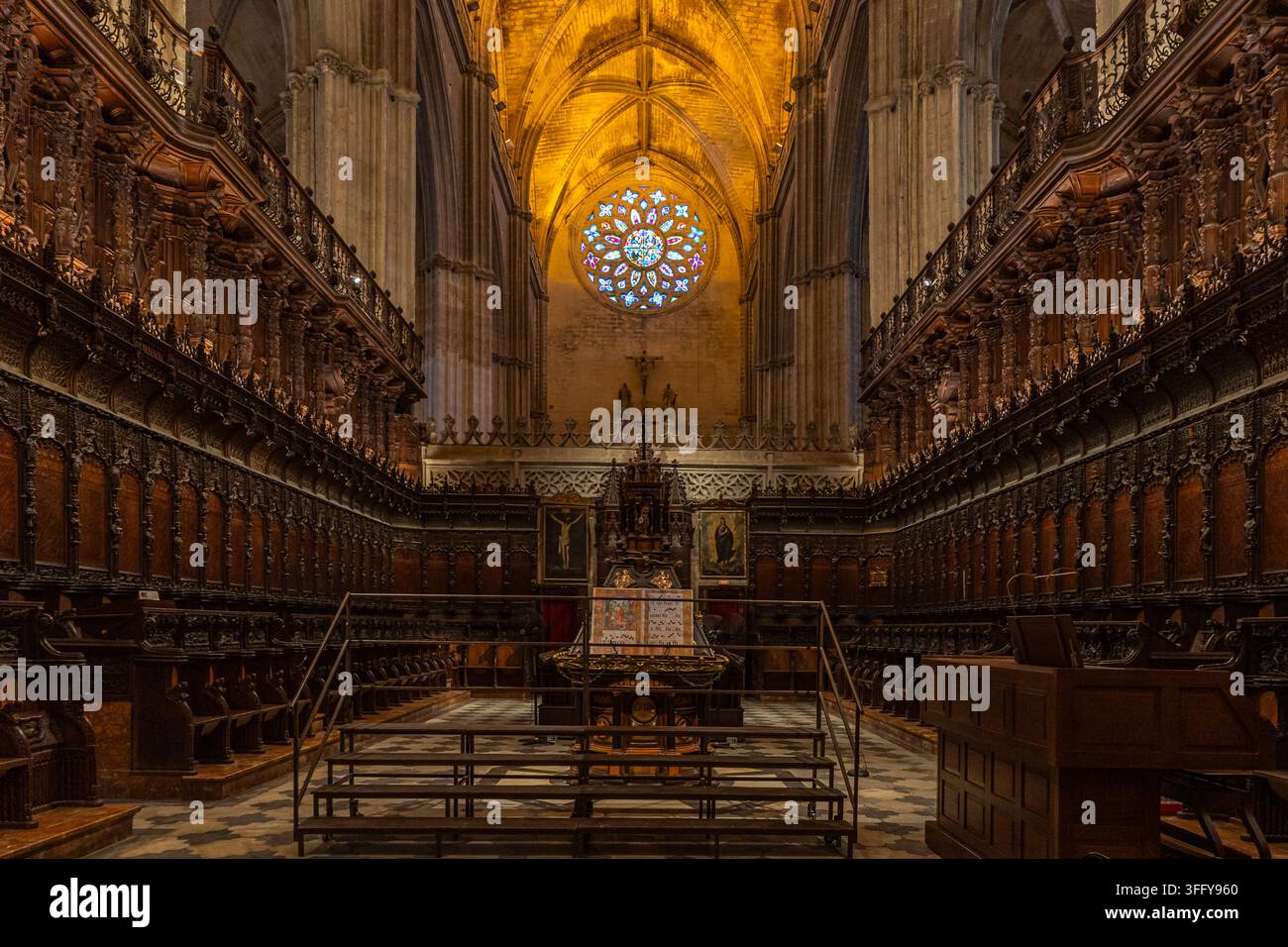 Schnitzereien an den Chorgestühlen bilden einen Blick auf ein farbenfrohes Rosettenfenster unter den hohen Gewölben der Kathedrale von Sevilla Stockfoto