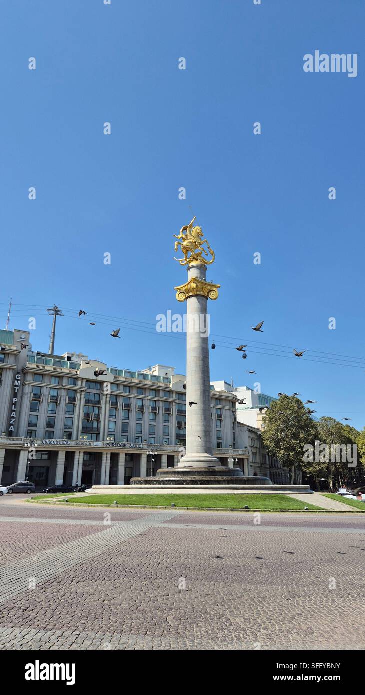 Statue des Heiligen Georg auf dem Freiheitsplatz in Tiflis, Georgien. Klare blaue Himmels mit einer Schar Vögel, die am Denkmal vorbeifliegen, und europäischer Architektur - Smartphone-aufgenommenes Stockfoto