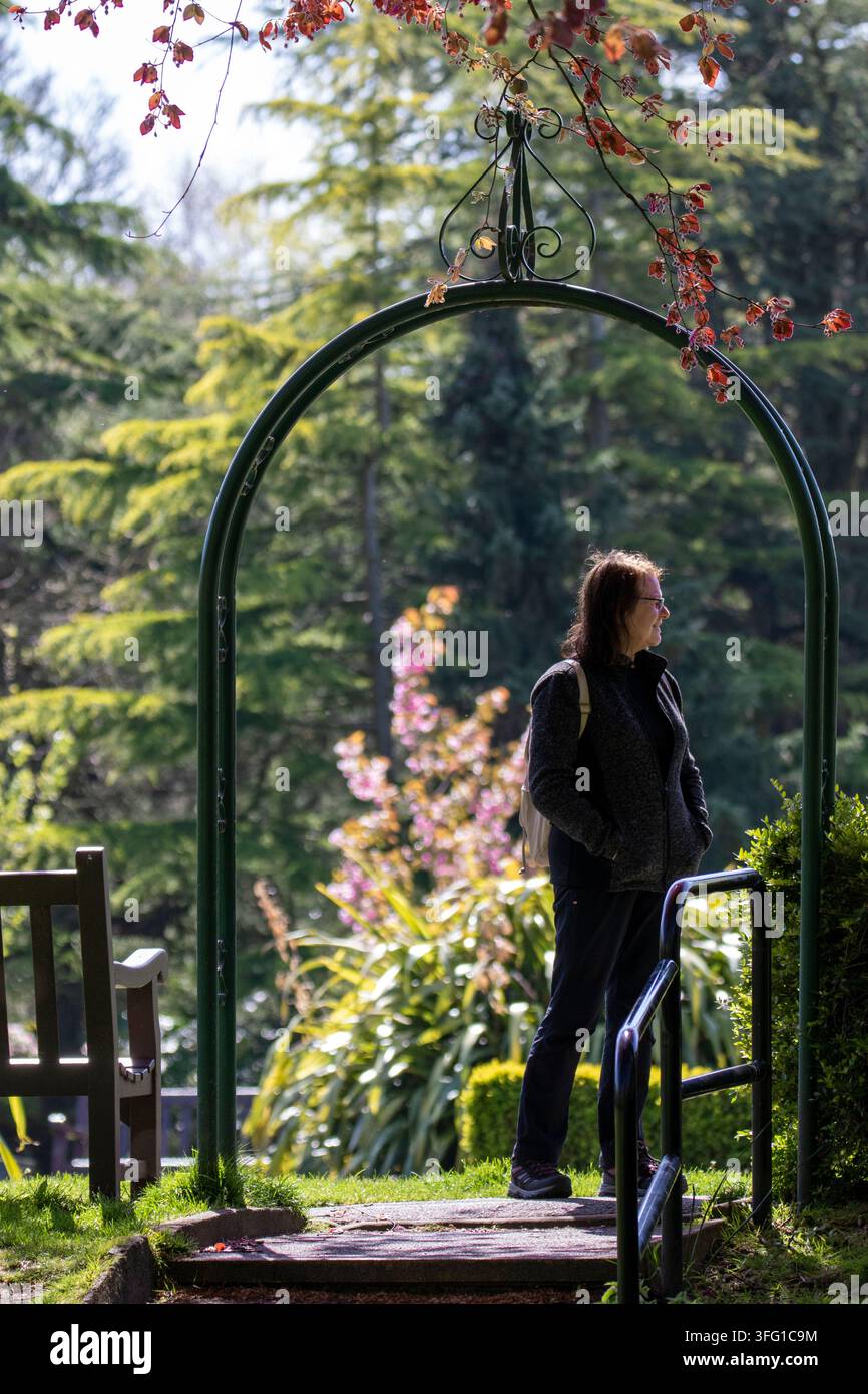 Eine Frau steht nachdenklich unter einem dekorativen metallenen Gartenbogen und blickt auf die malerischen Anlagen der Churchtown Botanic Gardens in Southport, UK. Stockfoto