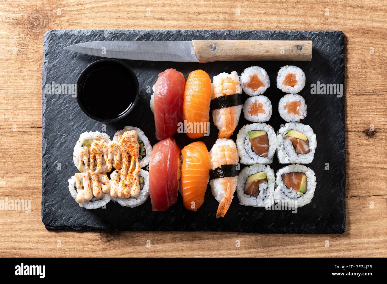 Verschiedenes japanisches Sushi auf einer schwarzen Schieferplatte serviert mit Lachs- und Thunfisch-Nigiri, Garnelen, Maki-Rollen, Sojasauce und einem Küchenmesser auf einem Holztisch. Stockfoto