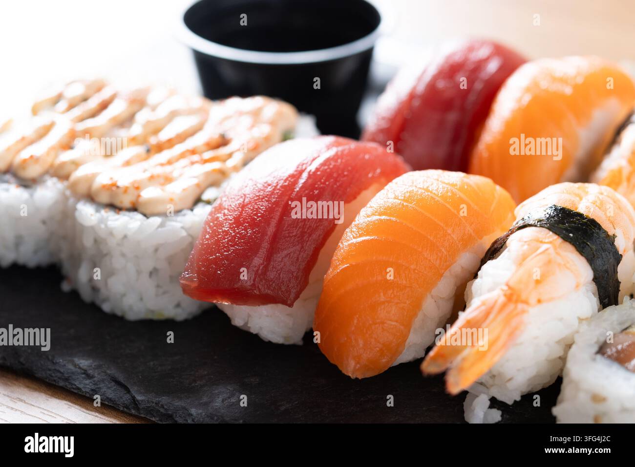 Verschiedenes japanisches Sushi auf einer schwarzen Schieferplatte serviert mit Lachs- und Thunfisch-Nigiri, Garnelen, Maki-Rollen, Sojasauce und einem Küchenmesser auf einem Holztisch. Stockfoto