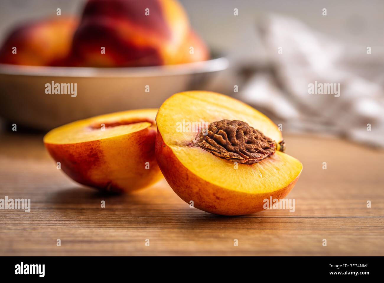 Eine Nahaufnahme einer reifen, halbierten Pfirsichfrucht, die das strukturierte Kernhaus und das saftige goldene Fruchtfleisch auf einem Holztisch zeigt, mit einem verschwommenen Schüssel voller Pfirsiche und weicher Küche Stockfoto