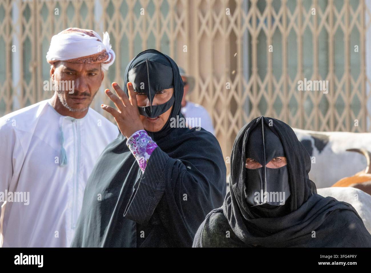 Omanischer Mann in einem weißen Turban steht in der Nähe von zwei Frauen, die traditionelle schwarze Gesichtsmasken tragen, auf dem Viehmarkt von Nizwa, Region Ad Dakhiliyah, Nizwa, O Stockfoto