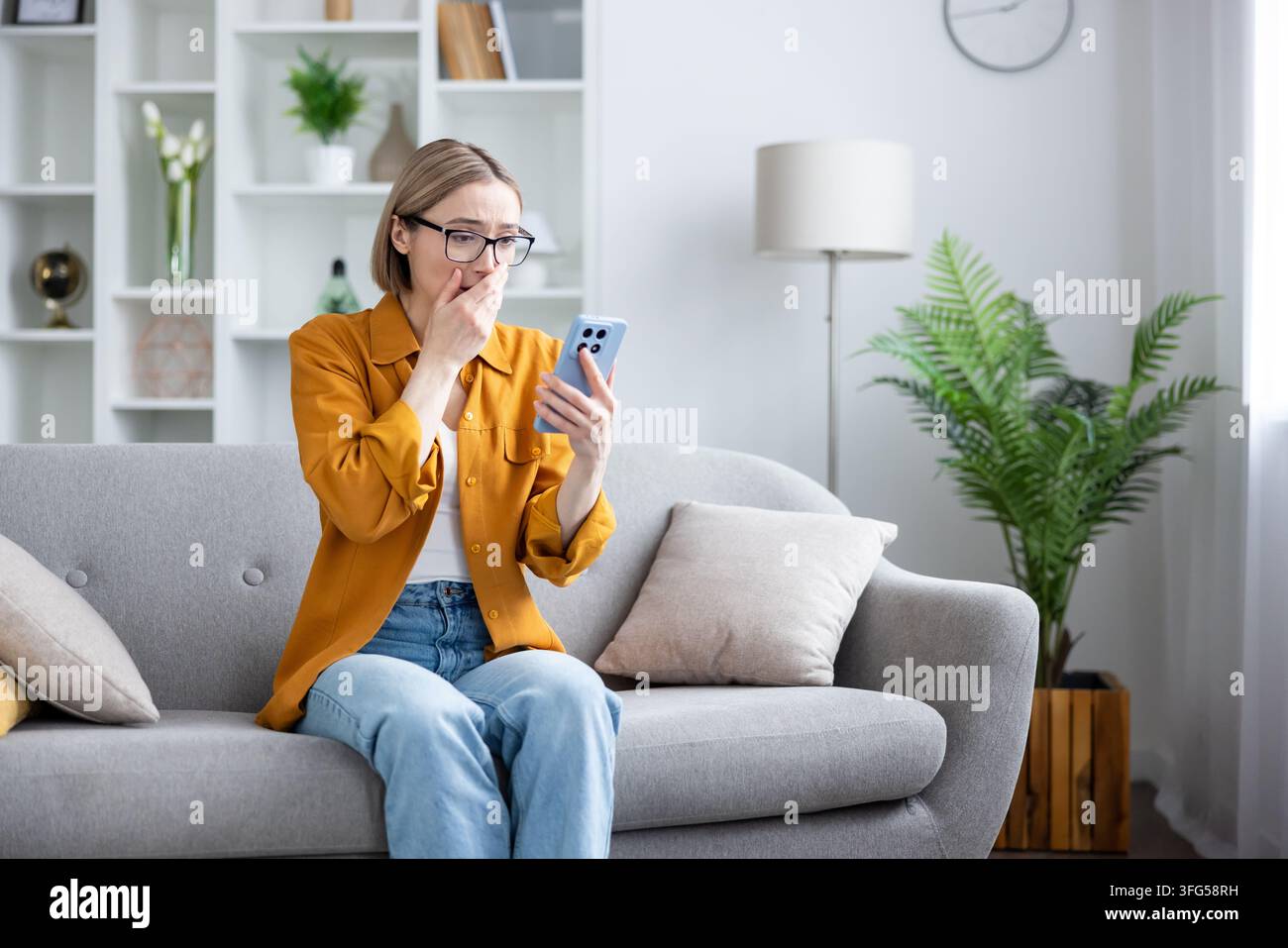 Frau, die auf einem Sofa in ihrem Wohnzimmer sitzt, schockiert und besorgt aussieht, während sie auf einen Handyd Bildschirm schaut, und Angst und emotionale Not über die Empfang von besorgniserregenden Informationen ausdrückt Stockfoto