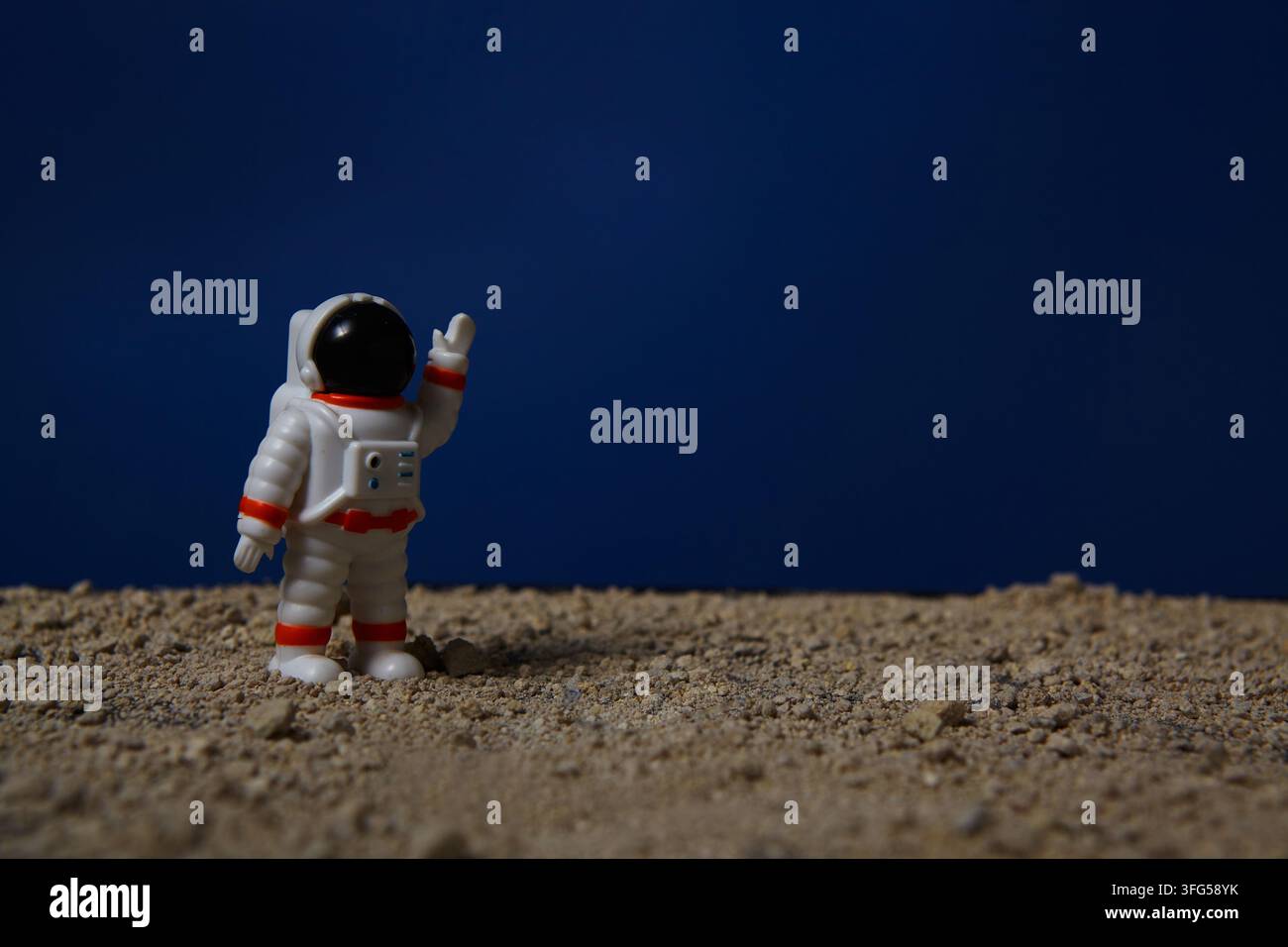 Ein Miniatur-Astronauten-Spielzeug steht auf einer felsigen, sandigen Oberfläche und hebt eine Hand vor einem tiefblauen Hintergrund. Stockfoto