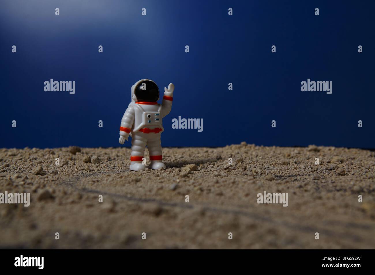 Ein Miniatur-Astronauten-Spielzeug steht auf einer felsigen, sandigen Oberfläche und hebt eine Hand vor einem tiefblauen Hintergrund. Stockfoto