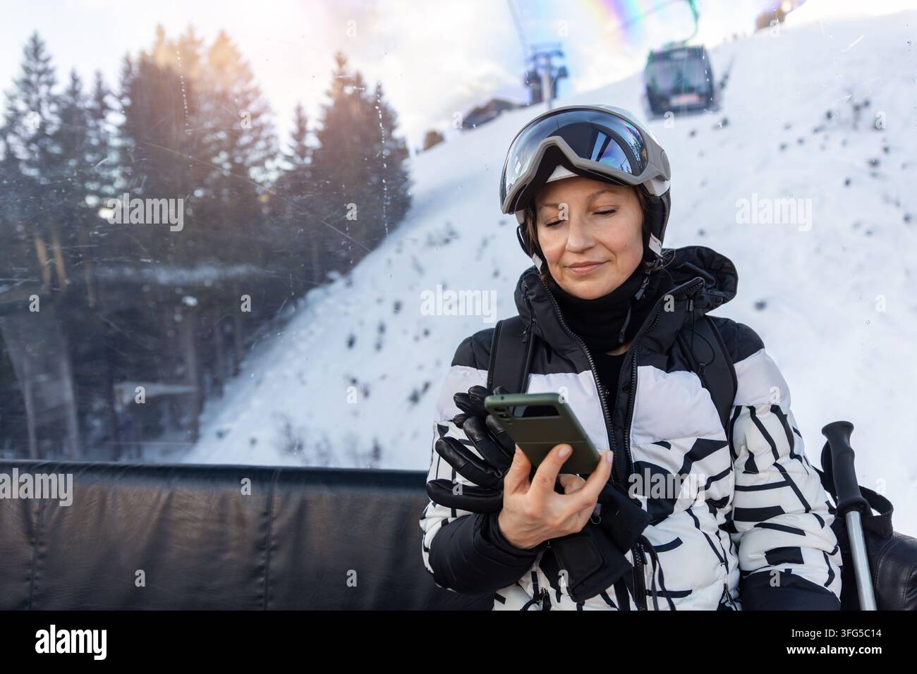 Frau mit Skihelm nutzt Smartphone in der Gondel beim Fahren durch einen verschneiten Alpenort. Wintersonne, Wald, Seilbahnen und Skigeräte Stockfoto
