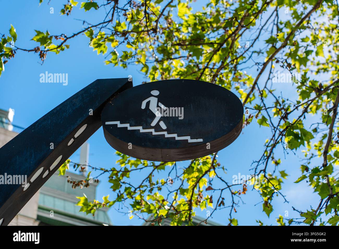 Rundes schwarzes Schild mit weißem Fußgänger-Treppen-Symbol vor blauem Himmel und grünen Blättern Stockfoto