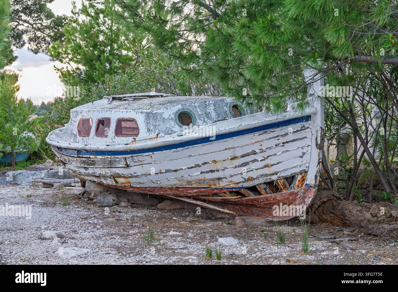 Ein verlassenes Holzboot mit abblätternder Farbe und verrostetem Rumpf ruht an einem felsigen Ufer zwischen Kiefern und vermittelt Verfall, Nostalgie und Küstenverlassenheit Stockfoto