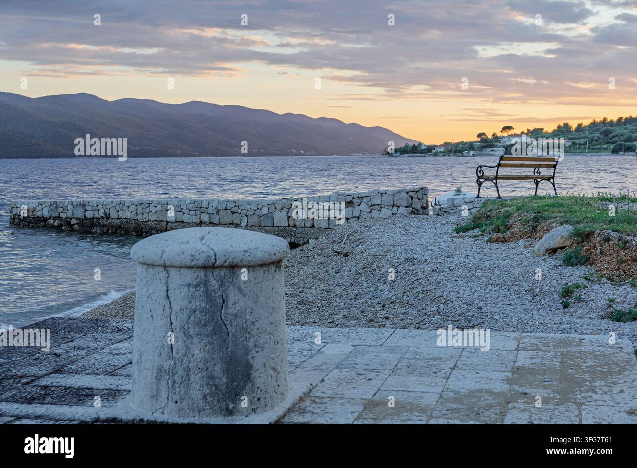 Eine leere Eisenbank blickt auf ein ruhiges Meer und entfernte Berge bei Sonnenuntergang, eingerahmt von einem Steinpier, einem kiesigen Ufer und einem weichen Pastellhimmel – eine friedliche Küste Stockfoto