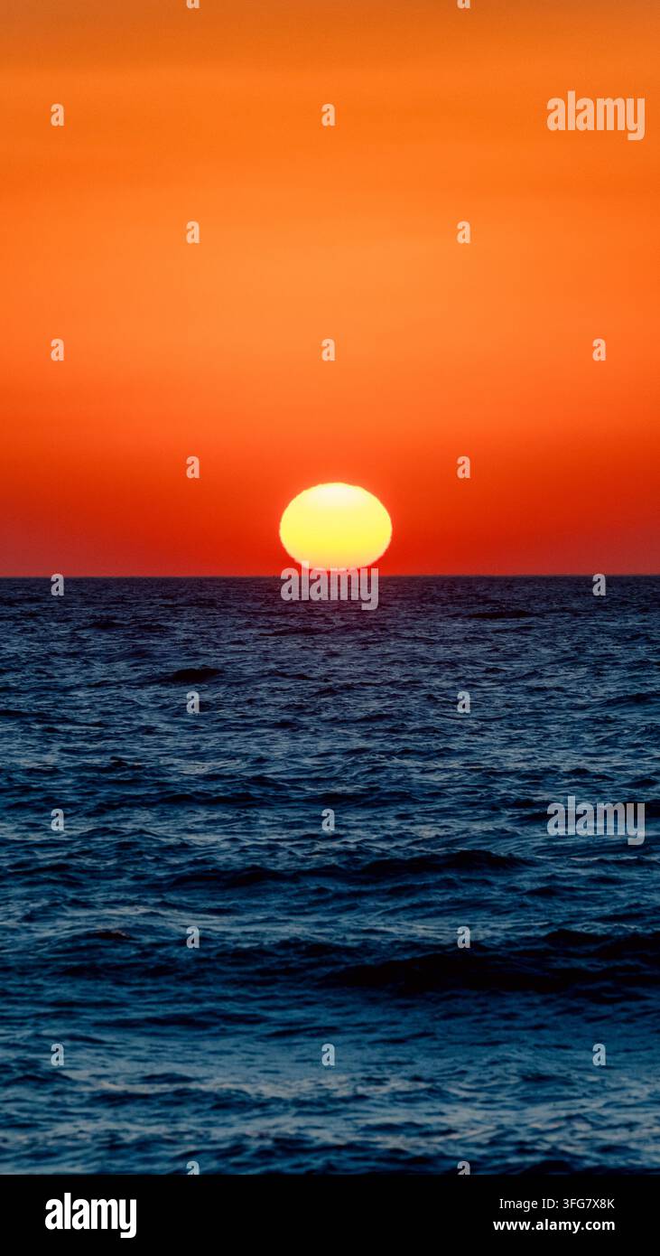 Helle Sonne, die sich direkt über dem Meereshorizont unter einem intensiven orangefarbenen Himmel setzt, wobei dunkelblaue Meereswellen starke warme und kalte Farbkontraste erzeugen Stockfoto