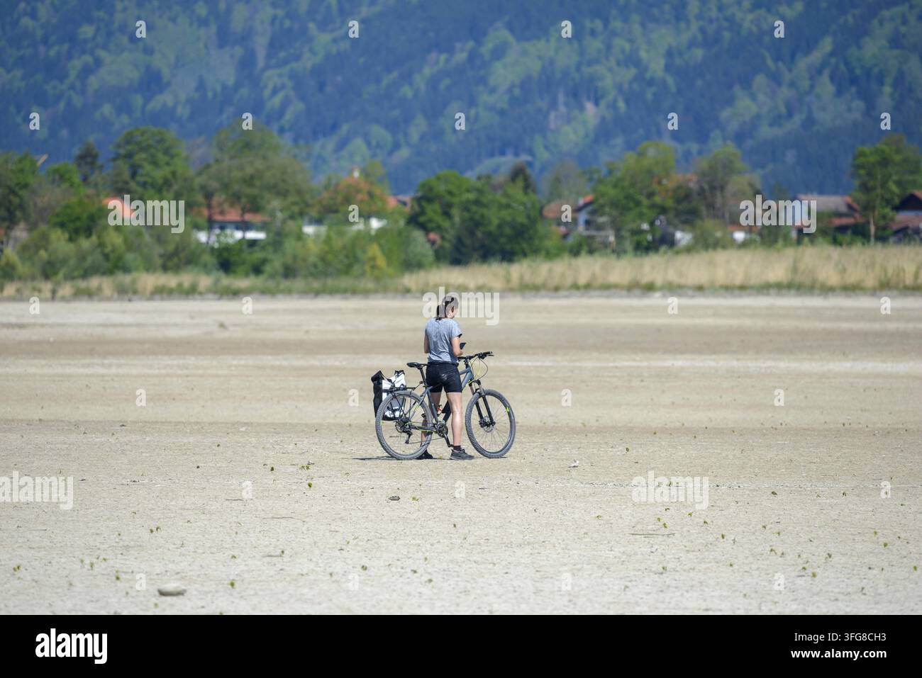 Frau mit Mobiltelefon, Fahrrad auf rissigem, trockenem Boden, wasserloser Forggensee im Frühling, nahe Fuessen, Ostallgäu, Bayern, Deutschland Stockfoto